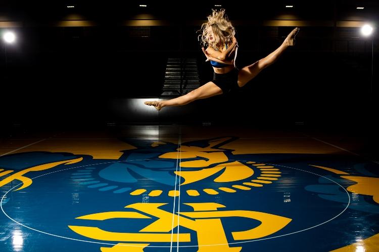 一个舞者以双腿劈叉的姿势在空中跳跃. 在她的下面是画在斯温尼篮球场地板上的小房间.