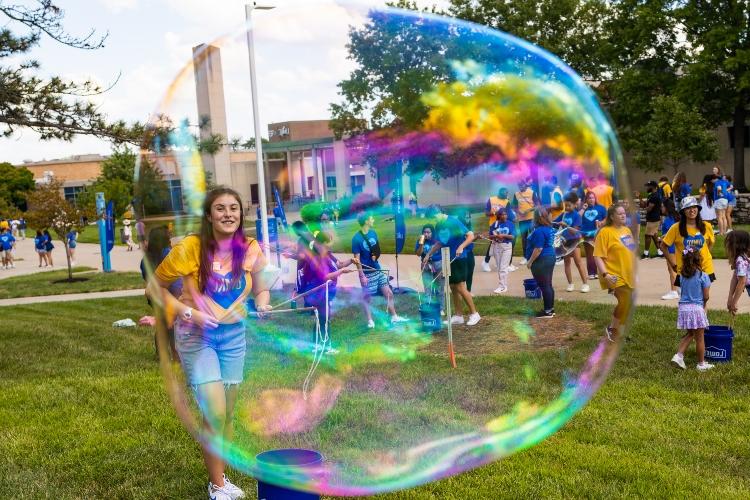 前景是一个半透明的气泡，后面是一个学生, 拿着泡泡棒微笑着走在阳光明媚的大学人行道上