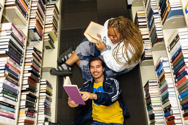 这张照片拍摄的是坐在图书馆两排书中间的两个学生. 一个坐在地板上看书，而另一个则把头靠在膝盖上看书.
